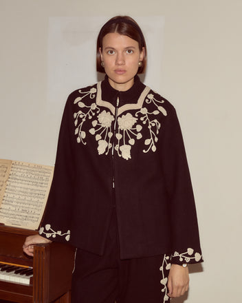 A woman in the Snowdrop Appliqué Jacket stands by a piano with sheet music, the elegant floral embroidery enhancing the charm of her swing silhouette wool outerwear.