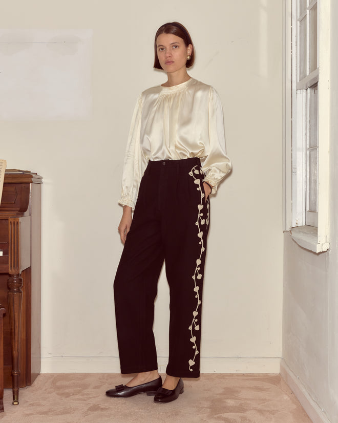 A woman stands indoors wearing black Snowdrop Appliqué Murray Trousers with floral embroidery on the sides and a white satin blouse.