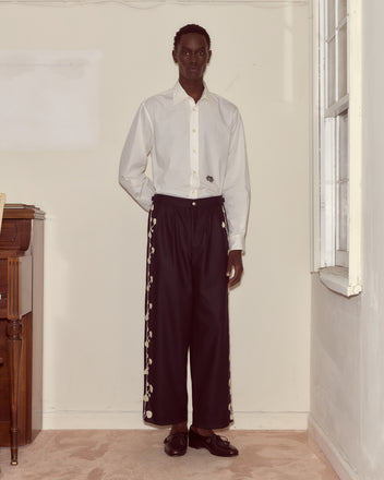 A person stands indoors wearing a white shirt and Snowdrop Appliqué Trousers in navy, featuring side embroidery.