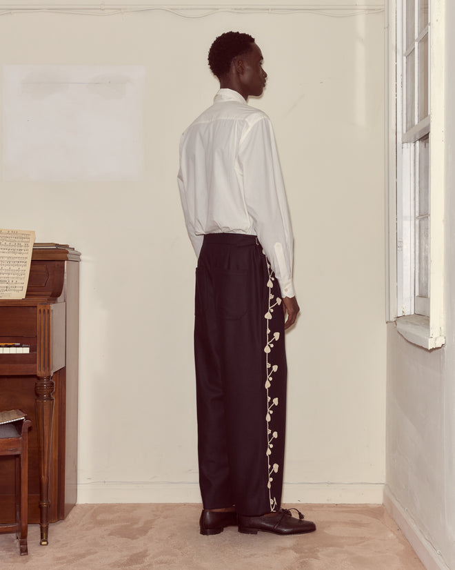 A man stands by a window, facing a piano, wearing black Snowdrop Appliqué Trousers and a white shirt.