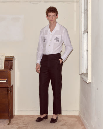 A man in a white shirt and Snowdrop Linen Side-Buckle Trousers stands indoors beside a piano and a window.