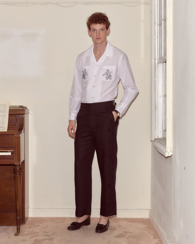 A man in a white shirt and Snowdrop Linen Side-Buckle Trousers stands indoors beside a piano and a window.