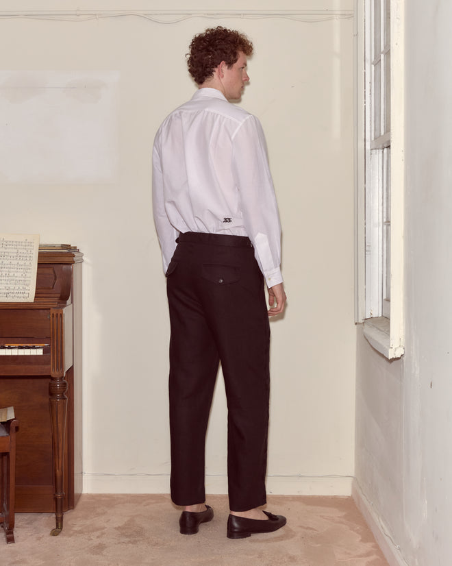 A man in a white shirt and Snowdrop Linen Side-Buckle Trousers stands facing away in a room with a piano and a window.
