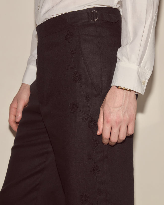 Model wears Snowdrop Linen Side-Buckle Trousers in black with subtle embroidery, paired with a white shirt, arm resting at their side.