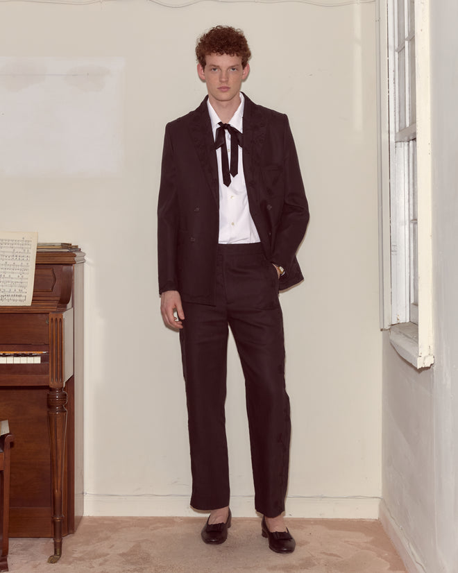Wearing the Snowdrop Linen Suit Jacket and a bolo tie, a person stands by a piano in a room with cream-colored walls.