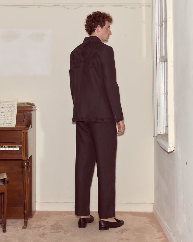 A man wearing the Snowdrop Linen Suit Jacket stands with his back to the camera beside a piano in a softly lit room.