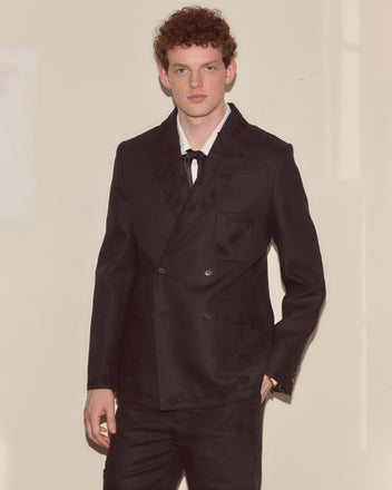 A young man with curly hair wears the Snowdrop Linen Suit Jacket, which features hand-crocheted floral appliqué, standing against a plain, light-colored wall.