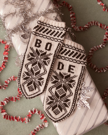 Snowflake Jacquard Mittens crafted from knitted merino wool, featuring the iconic BODE logo, rest elegantly on a wrapped gift adorned with silver tinsel and decorative trim.