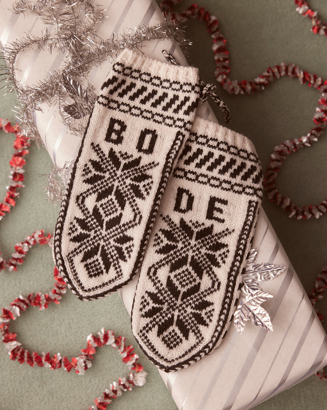 Snowflake Jacquard Mittens crafted from knitted merino wool, featuring the iconic BODE logo, rest elegantly on a wrapped gift adorned with silver tinsel and decorative trim.