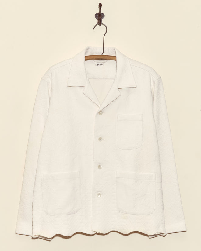 The Snow Trellis Overshirt - L, a unique white quilted shirt with three front pockets, hangs on a wooden hanger against a beige wall.