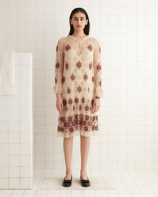 A woman in the Solarium Dress, a 1930s-inspired piece featuring a beige and red pattern, stands gracefully in a white tiled room.