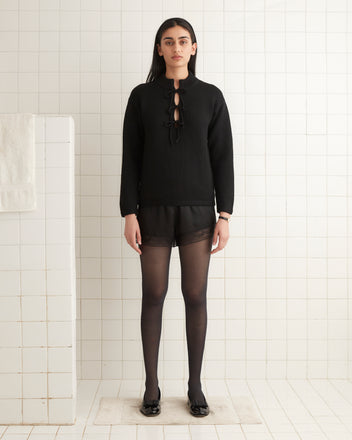 A person wearing the Solid Front Tie Pullover in black, oversized style stands in a bright tiled room with a towel on a rack.