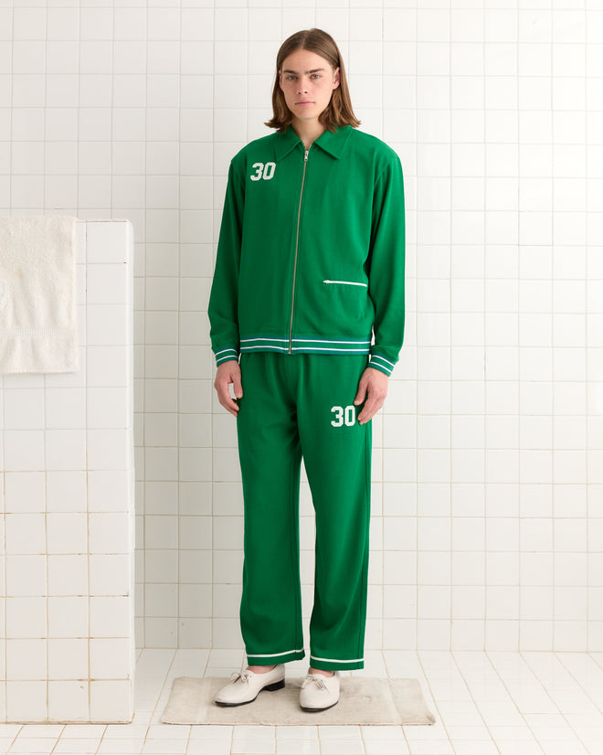 In a white tiled room, someone in a green "30" Sophomore Jacket stands next to a towel, capturing the classic charm of vintage jerseys. Their nostalgic style and heavyweight fabric evoke memories of a bygone era.