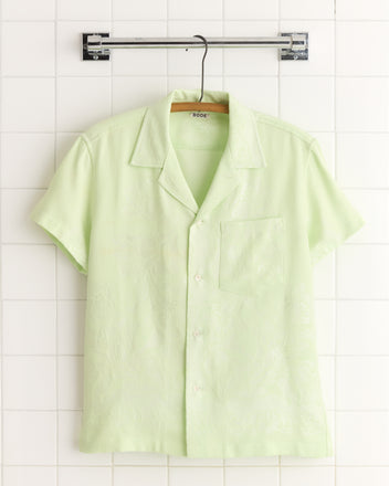The Sorbet Crystal Shirt (medium) in light green damask, short-sleeved with a collar, made in New York, hangs on a hanger against a tiled wall.