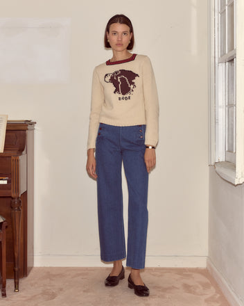 Woman stands indoors wearing a Spaniel Sweater crafted from a cozy cotton merino blend, paired with blue jeans and black shoes.