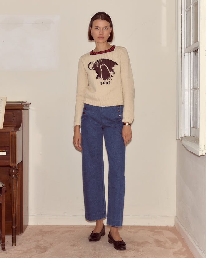 Woman stands indoors wearing a Spaniel Sweater crafted from a cozy cotton merino blend, paired with blue jeans and black shoes.