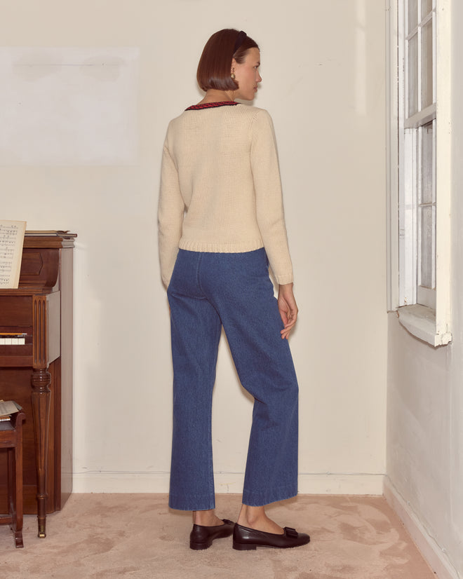 A woman stands indoors facing a wall, wearing a cream Spaniel Sweater, blue pants, and brown loafers.