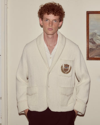 A man with curly red hair stands indoors wearing the Sportsman Jacket, a cream waffle cotton style with an embroidered crest inspired by classic men’s robes.