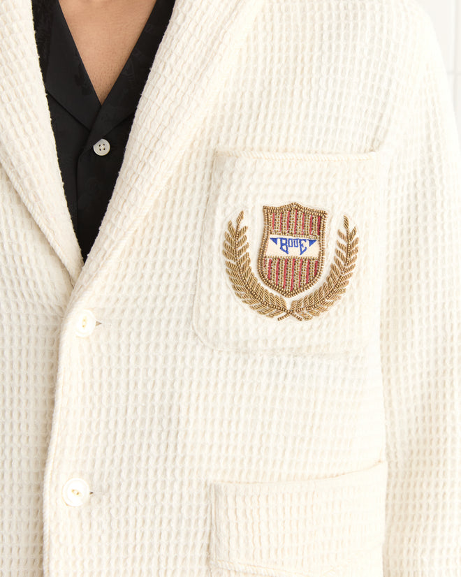 Close-up of the Sportsman Jacket, a cream textured blazer made from waffle cotton, adorned with an embroidered bullion crest and laurel embroidery on the pocket, paired with a black shirt.