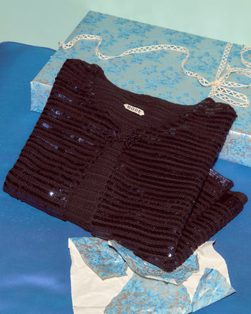 The Sequin Spotlight Swing Cardigan in Black/Blue, featuring shimmering sequins and a “BODE” label, rests on blue satin next to a blue floral box and tissue—luxurious knitwear with standout style.