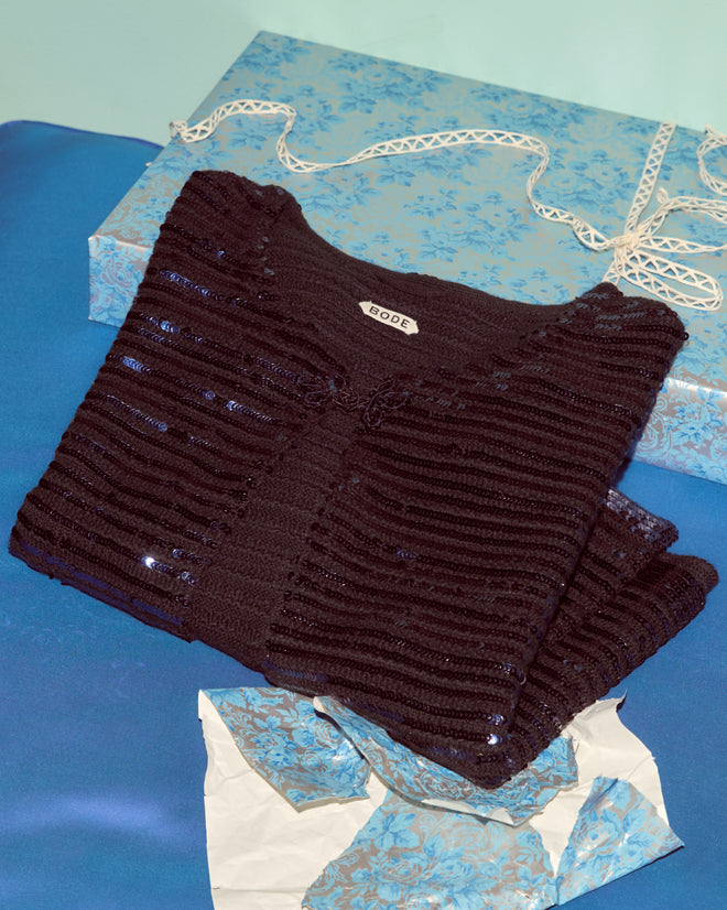 The Sequin Spotlight Swing Cardigan in Black/Blue, featuring shimmering sequins and a “BODE” label, rests on blue satin next to a blue floral box and tissue—luxurious knitwear with standout style.