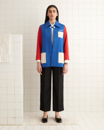 A person wearing the vibrant Squad Jacket, featuring blue and red colorblocking with chain-stitched details, stands in a white tiled room, paired with black pants. The eye-catching design adds a stylish touch to their ensemble.