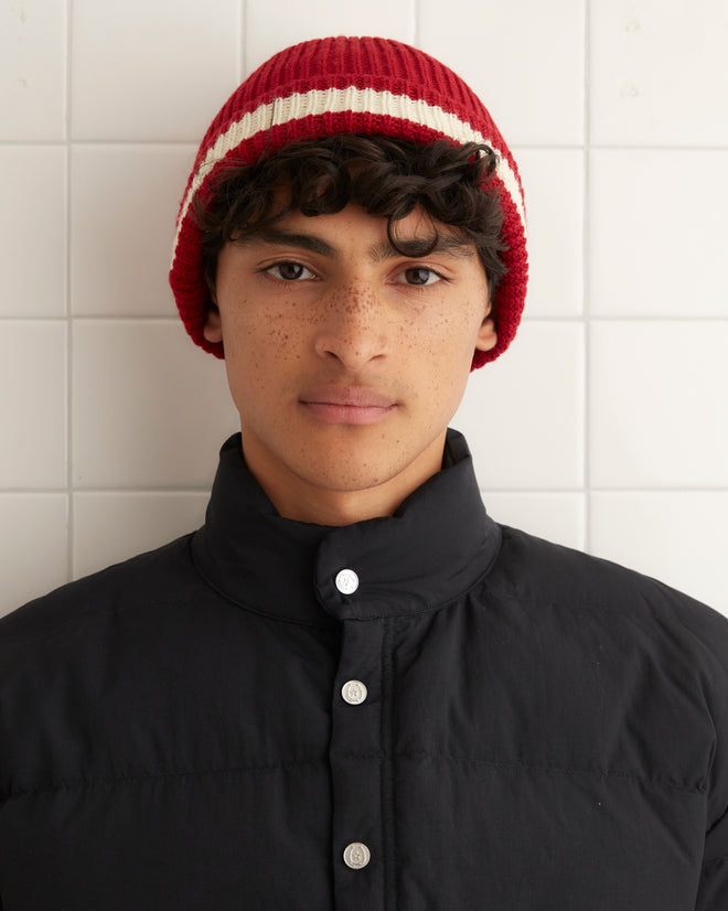 The Pom Hat - Red showcases a charming vintage knit design in red, accented with a cream stripe and a playful pom-pom, all set against a tiled background. Crafted in Peru, this comfortable accessory is made with premium merino wool for unparalleled style and coziness.