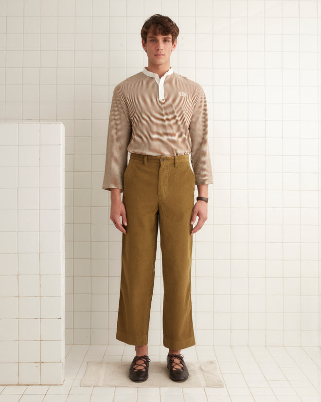 The Corduroy Standard Trousers in olive green, featuring midweight fabric and an adjustable back buckle, hang on a wooden hanger against a white wall, evoking the timeless style of 1940s French workwear.