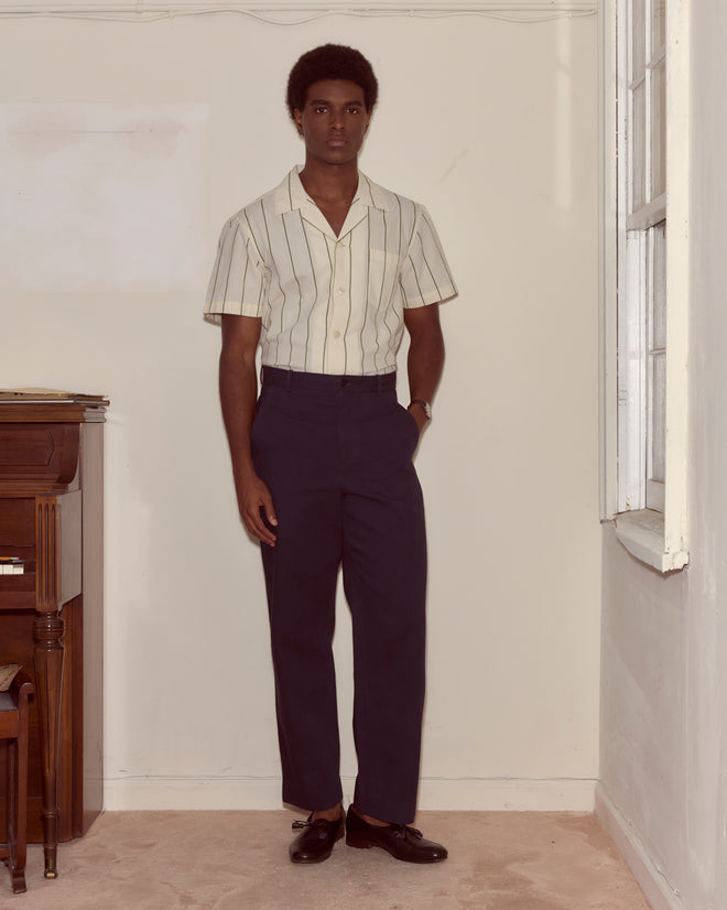 A man stands indoors beside a piano wearing a striped short-sleeve shirt, Standard Trousers - Navy, and loafers.
