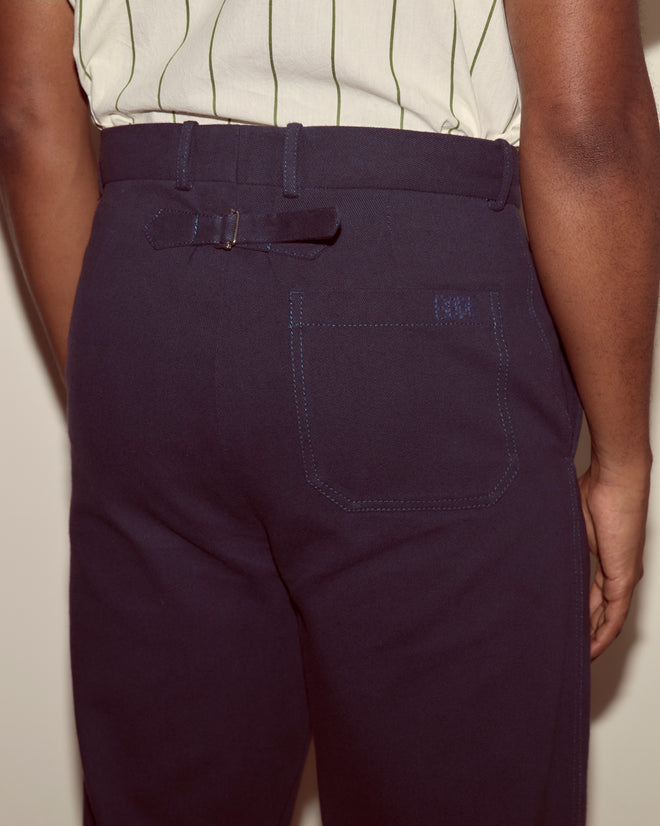 A person is seen from behind wearing the Standard Trousers - Navy, featuring a back pocket and belt, with a striped shirt partially visible.