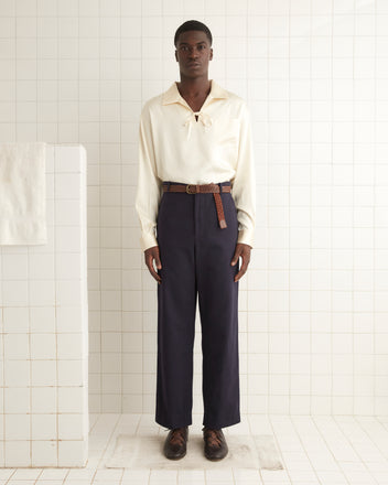 A man stands in a tiled room wearing a cream long-sleeve shirt with a tie at the neck, French workwear trousers named 