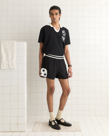 A person in a black sports outfit, paired with white socks and shoes, stands elegantly in a tiled room, sporting the shimmering Star Field Shorts - Black.