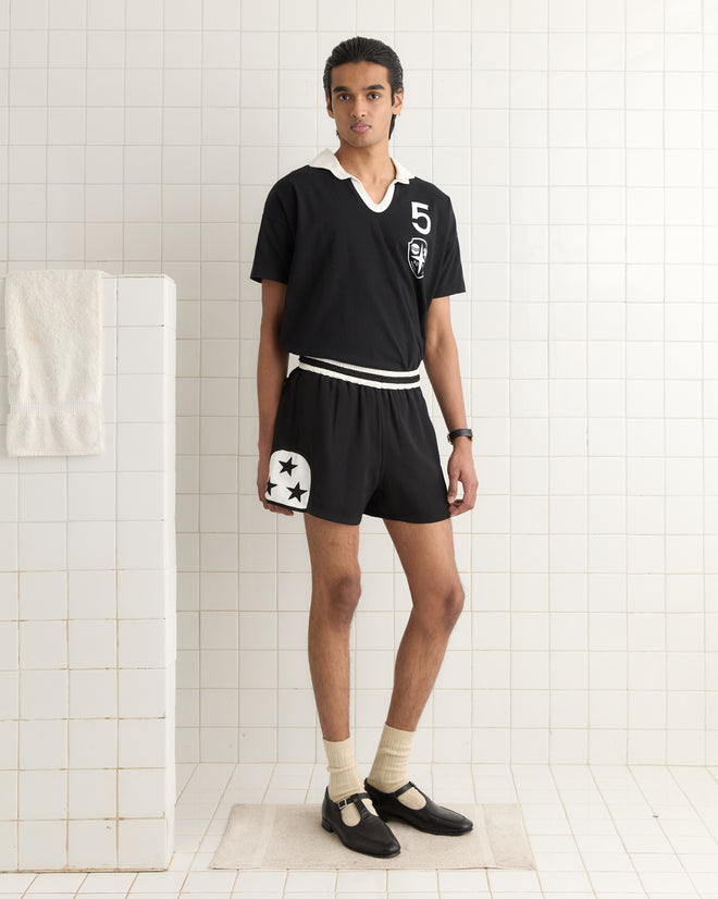 A person in a black sports outfit, paired with white socks and shoes, stands elegantly in a tiled room, sporting the shimmering Star Field Shorts - Black.