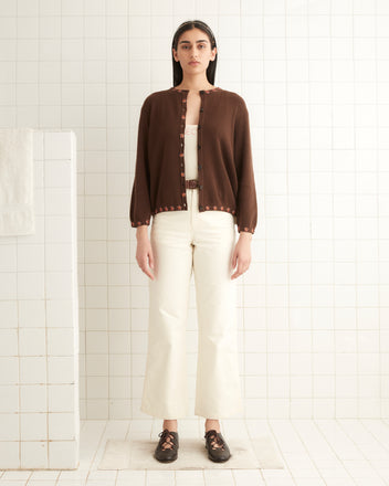 A woman in a brown Starflower Beaded Cardigan featuring intricate beaded flower appliqués, paired with white pants, stands in a tiled room, gazing forward.