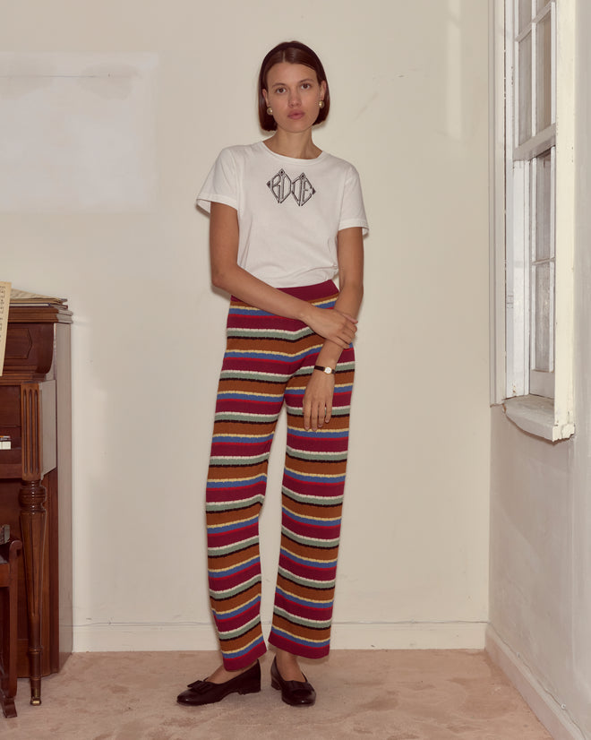 A woman wearing Starkey Pants and a white t-shirt stands by a window indoors, next to a piano and sheet music.
