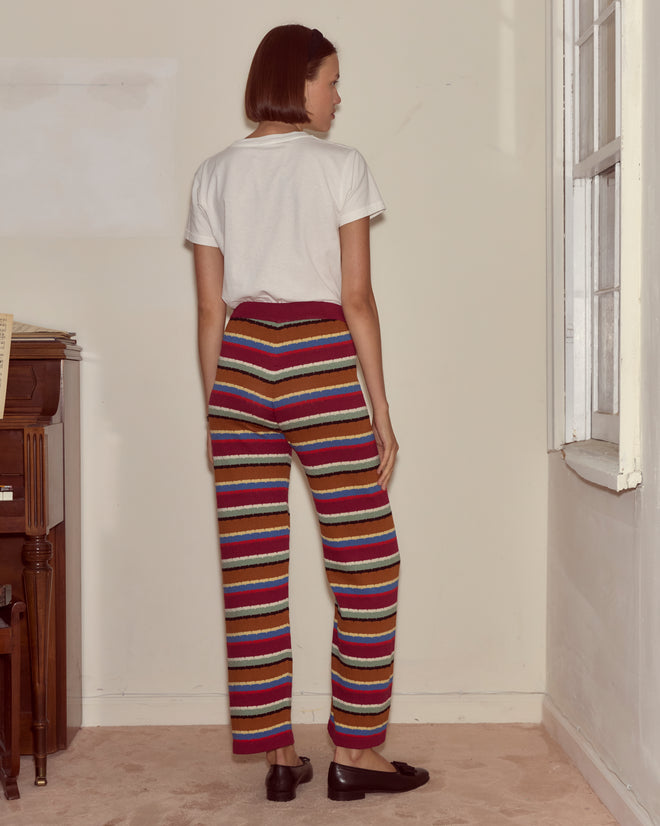 A person stands indoors by a window, wearing a white shirt and the Starkey Pants—colorful wide-leg striped trousers.