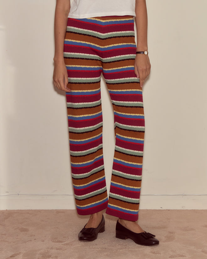 Woman wearing Starkey Pants—colorful wide-leg striped style—with a white top and brown shoes; beige carpet and plain wall in the background.