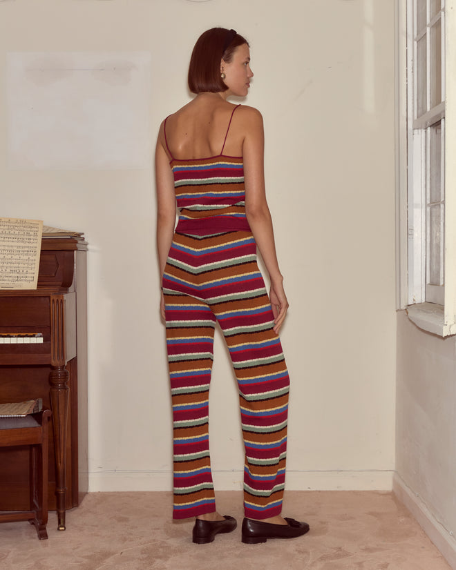 Woman with short hair wearing the Starkey Tank and a striped jumpsuit stands by a piano, facing away, near a window in a bright room.