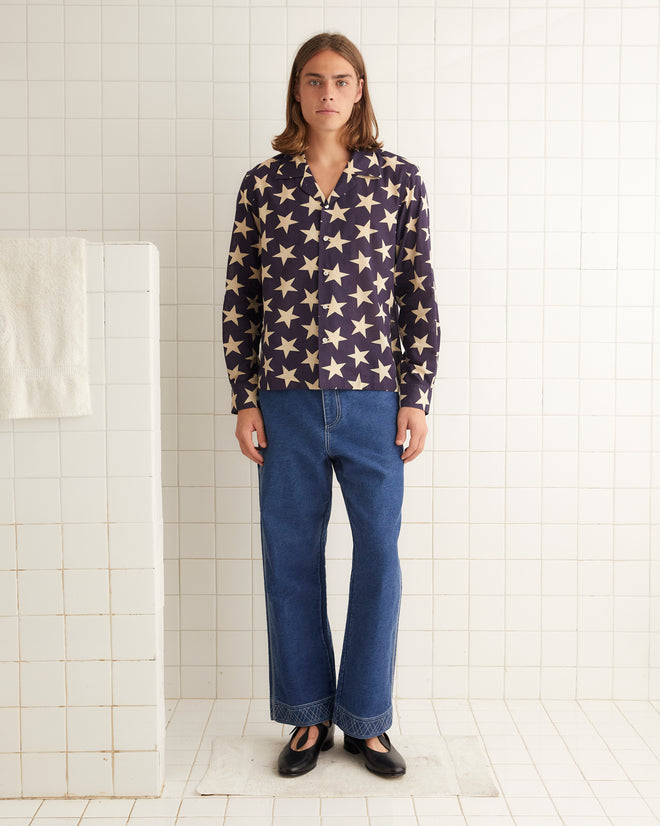 A person stands in front of blue curtains, wearing a 100% cotton Starry Long Sleeve Shirt adorned with intricate embroidered Bode details and paired with white pants, with one arm resting on their shoulder.