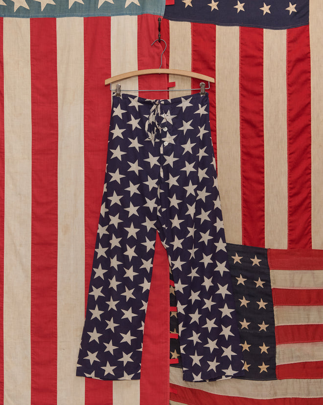 Starry Pajama Pants, crafted from 100% cotton and featuring a blue star pattern, hang on a wooden hanger in front of an American flag backdrop.