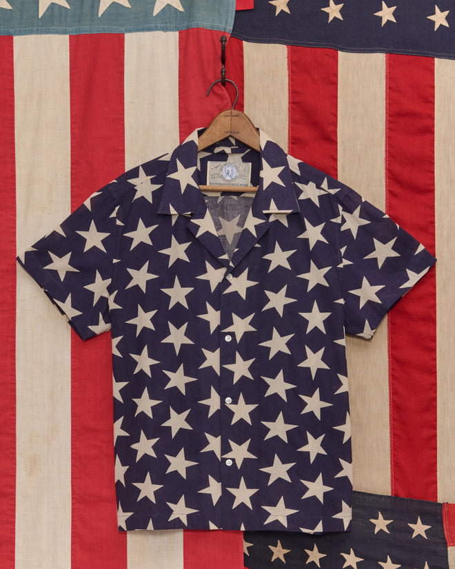 The Starry Short Sleeve Shirt features a vintage-inspired star pattern in navy and white, displayed on a wooden hanger against an American flag backdrop.