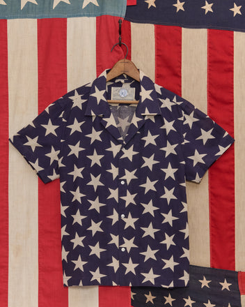 The Starry Short Sleeve Shirt features a vintage-inspired star pattern in navy and white, displayed on a wooden hanger against an American flag backdrop.