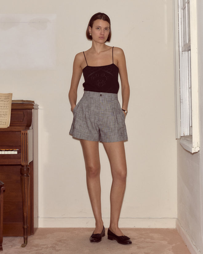 A woman in a black tank top and Static Weave Shorts stands indoors by a piano, hands in pockets.