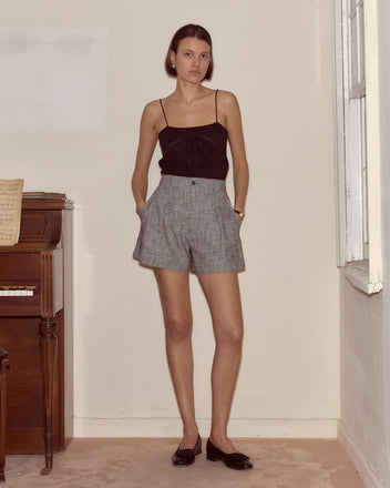 A woman in a black tank top and Static Weave Shorts stands indoors by a piano, hands in pockets.