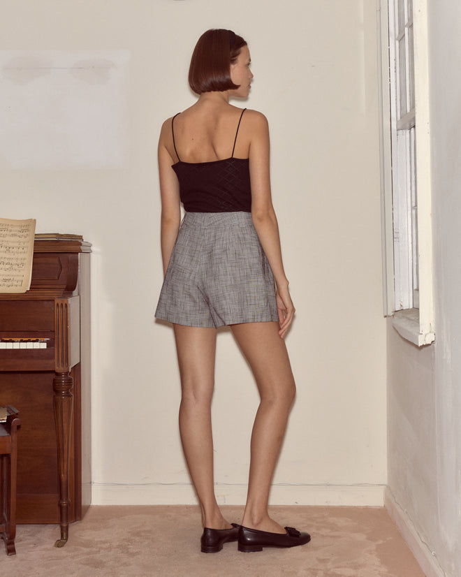 A woman with short hair, wearing a black top and Static Weave Shorts in gray, stands facing a wall beside a piano and window.
