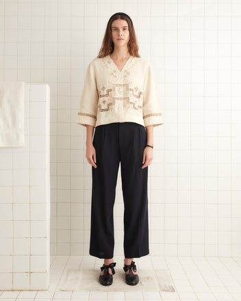 A person with long hair stands on a white tiled floor. They wear the Step Cutwork Overshirt, a cream blouse adorned with antique pearl buttons and intricate embroidery, paired with black pants and matching shoes. The background features white tiled walls and a partially seen towel.