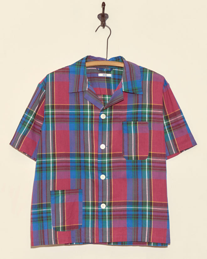 The Stone Fruit Plaid Shirt - S/M is a short-sleeve, button-up, deadstock shirt with a red, blue, yellow, and green plaid pattern, displayed hanging on a hook.