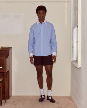 A person stands indoors wearing a Striped Poplin Shirt, black shorts, white socks, and black shoes.