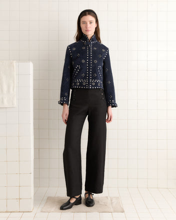 In a tiled room, a person is stylishly clad in the Studded Howard Jacket - Dark Navy and black pants, their hands resting by their sides.