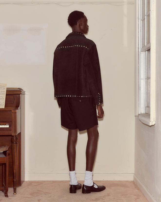 A person wearing the Studded Welding Jacket - Black, along with shorts, white socks, and black shoes, stands facing a beige wall near a piano.